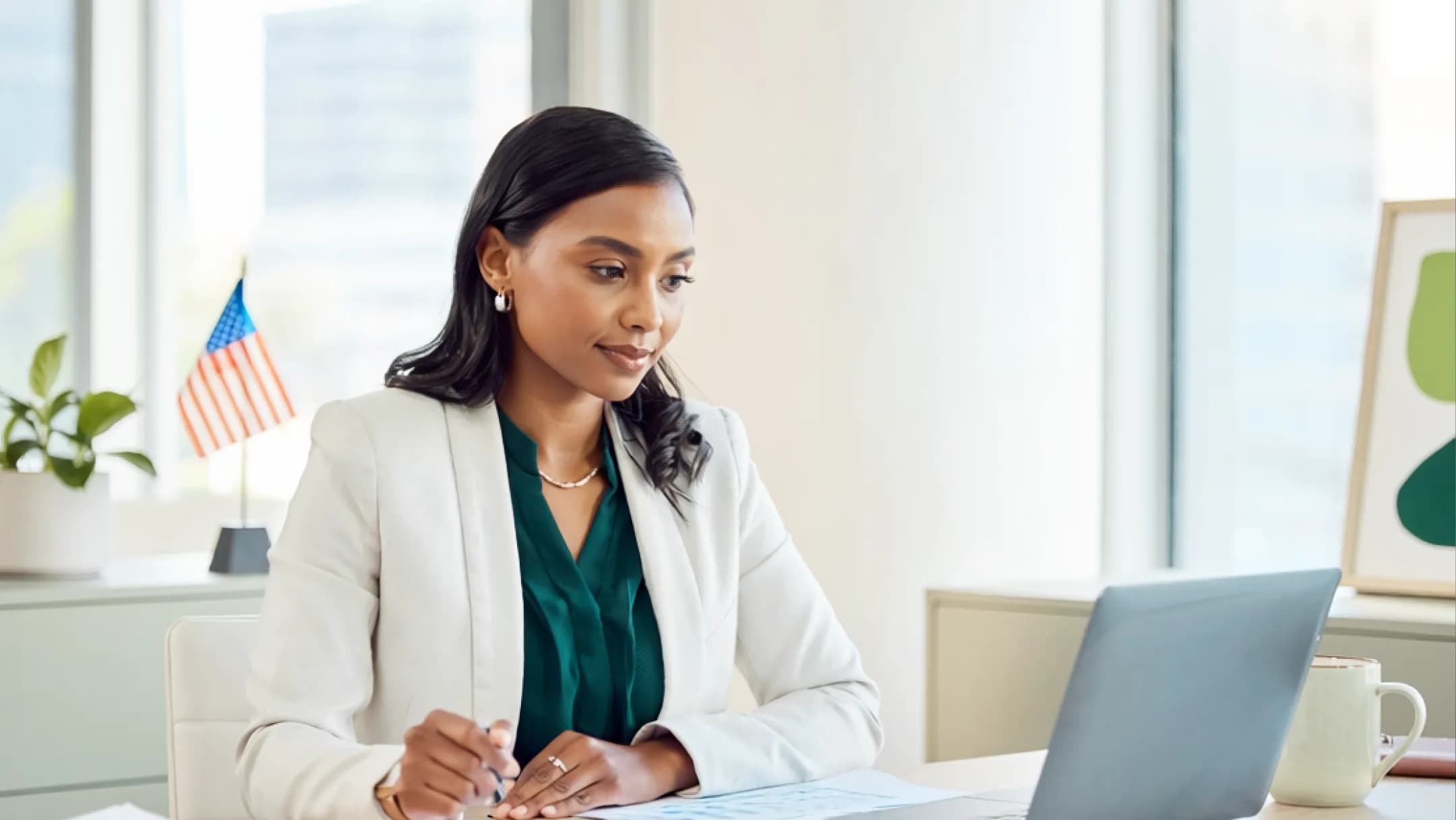 A businesswoman at a laptop beside a small U.S. flag in a bright office